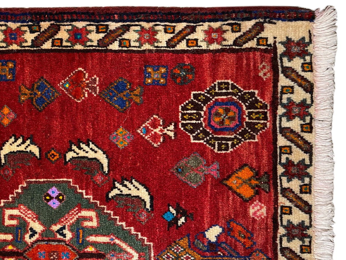Vista aproximada de um tapete Poshti de 58x57 cm: pelo vermelho com ornamentos e franja nas extremidades