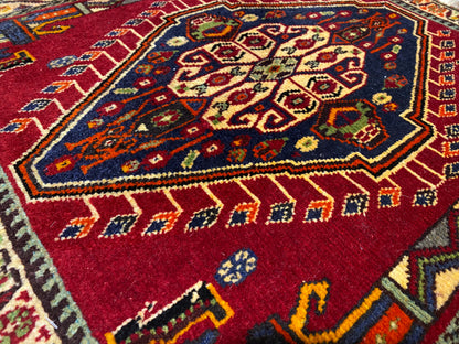 Close-up de um tapete Poshti (59×61 cm): padrão interior vermelho com ornamentos coloridos e moldura