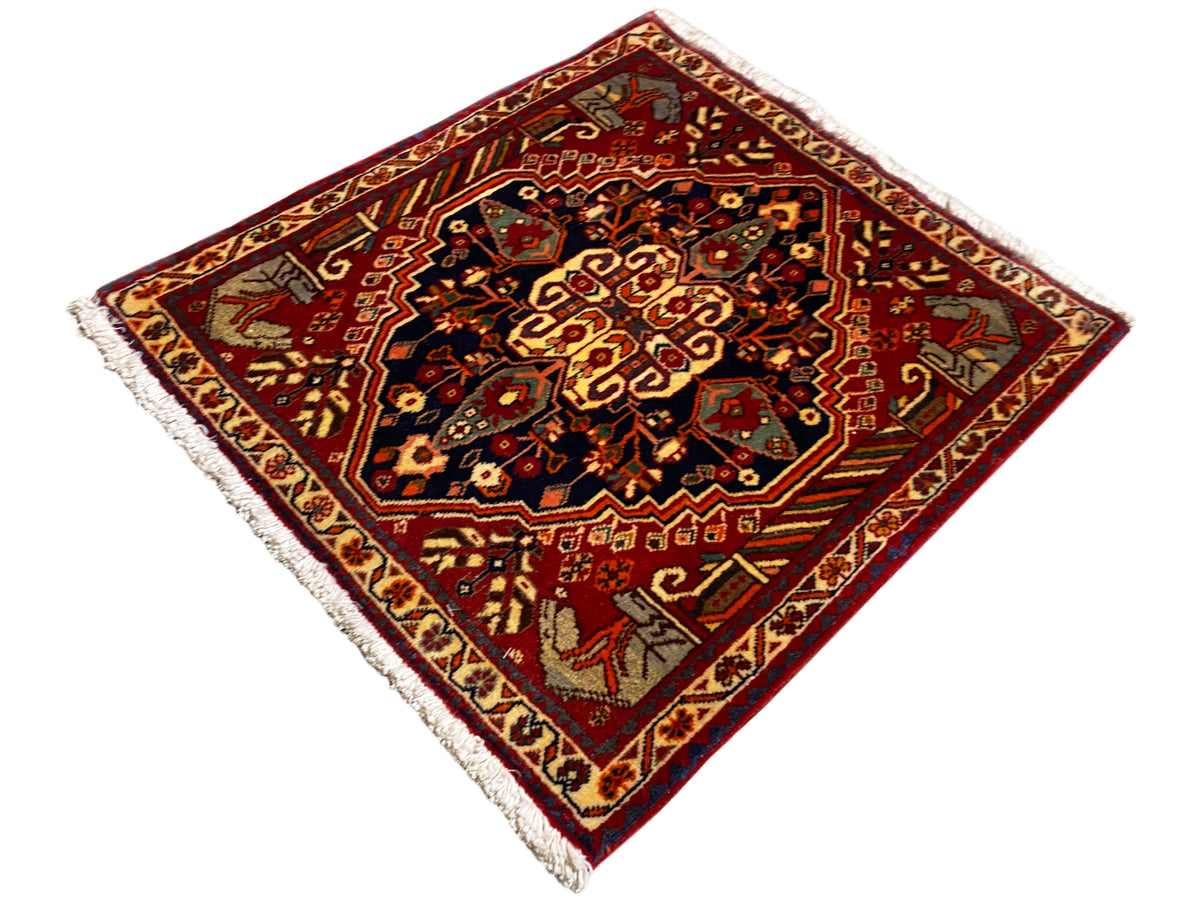 Tapete de 65x59 cm com fundo vermelho, ornamentos centrais escuros e franjas nas extremidades